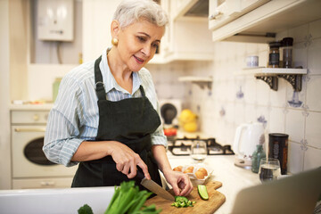 Attractive gray haired female pensioner in apron standing at kitchen table holding knife, cutting cucumber on board while making new vegetable meal, watching online video recipe on laptop