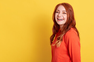 Laughing woman with happy facial expression looking away, wearing orange casual sweater, has good news, copy space for advertisement or promotions, ginger lady looks happy, against yellow wall.
