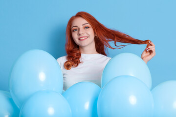 Obraz na płótnie Canvas Lovely ginger female flirts with someone and pulling her hair aside, wears white shirt, poses with inflated balloons against blue background. Birthday party, celebrating.