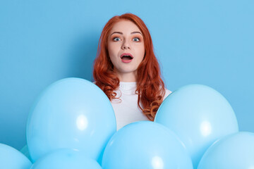 Surprised young red haired woman girl dresses white shirt, posing isolated on blue background. Birthday holiday party. Female with opened mouth hold air balloons, looks at camera with shock, forgot.