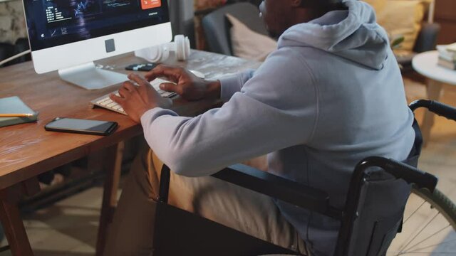 Midsection shot of disabled Afro-American man on wheelchair sitting at desk and working on computer at home