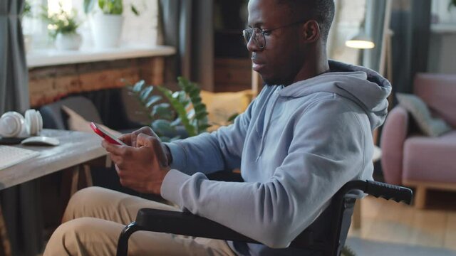 Zoom in shot of young disabled Afro-American man on wheelchair typing on smartphone and then looking at camera in living room at home