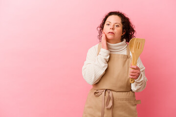 Woman with Down syndrome cooking at home isolated on pink background is saying a secret hot braking news and looking aside