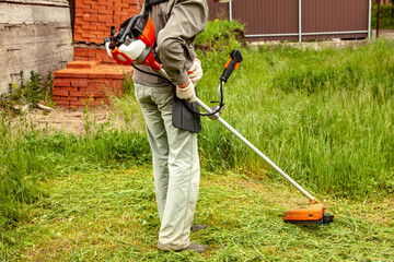 A man in work clothes mows tall grass with a trimmer in the garden