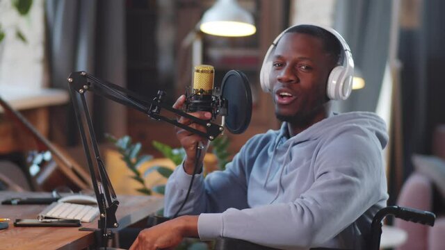 Young disabled Afro-American man on wheelchair in wireless headphones talking into microphone and posing for camera with smile while recording podcast in home studio