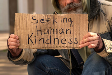 Senior man homeless beggar with a gray beard in a shabby clothes with a carboard sign sitting outdoors in city and asking for money donation. Sign on paperboard Seeking Human Kindness - obrazy, fototapety, plakaty
