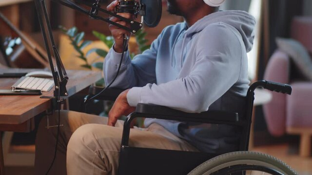 Tilt up shot of young disabled Afro-American man on wheelchair in wireless headphones speaking into microphone while recording podcast in home studio