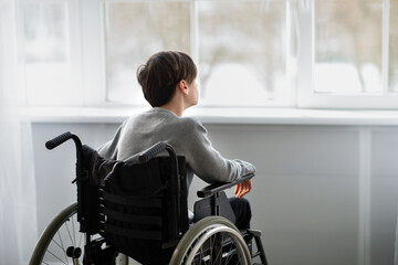 Back view of disabled teenager in wheelchair looking out window, having depression at home, copy...