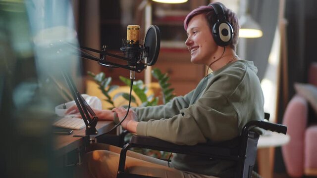 Tilt up shot of young disabled woman on wheelchair in wireless headphones talking into microphone and using computer while recording podcast in home studio