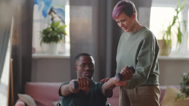 Young disabled Afro-American man on wheelchair doing arm exercises with dumbbells with assistance of female caregiver at home