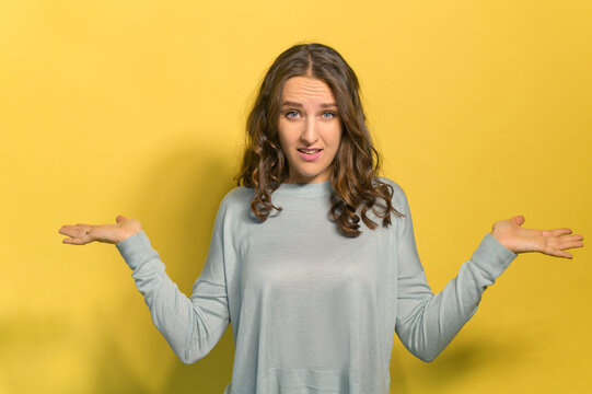Uncertainty concept. A young curly brunette woman spreads her arms and shrugs shoulders, does not know what to do, a female lost in doubts, studio shot isolated on yellow
