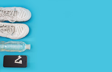 Healthy lifestyle and sport concept. White sneakers, bottle of water, smartphone with headphones on a blue background. Copy space.