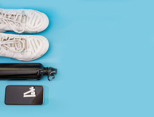 Healthy lifestyle and sport concept. White sneakers, bottle of water, smartphone with headphones on a blue background. Copy space.