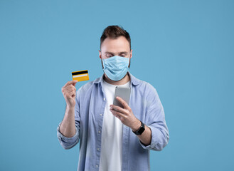 Online shopping during covid. Young guy in face mask purchasing goods on web, holding cellphone and credit card