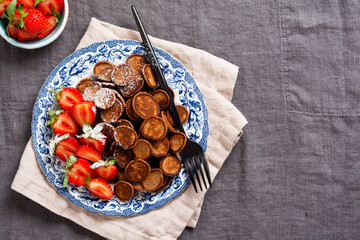 Mini chocolate pancake cereal with strawberries for breakfast on gray old textile tablecloth. Trendy home breakfast with tiny pancakes. Top view