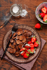 Mini chocolate pancake cereal with strawberries for breakfast on old wooden table. Trendy home breakfast with tiny pancakes. Top view