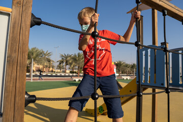 Obraz premium Young boy in a COVID-19 mask climbing and playing on the playground in the summer sunshine for recreational fun during pandemic.