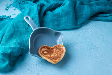 Pancake in the shape of a heart in a flying pan, turquoise fabric on a blue background. Horizontal orientation. 