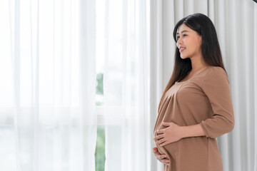 Asian happy pregnant woman is standing beside windows and touching her belly. pregnancy, motherhood, people and expectation concept