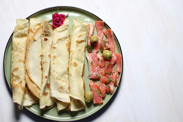 Pancakes on green plate with salty salmon slices, olives, rose flower on white table