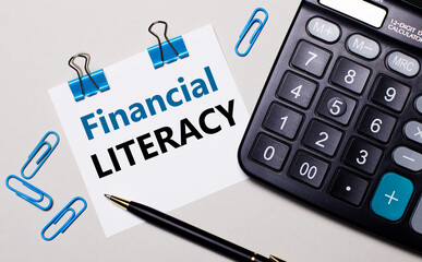 On a light background, a calculator, a pen, blue paper clips and a sheet of paper with the text FINANCIAL LITERACY. View from above