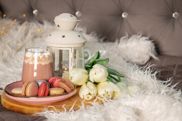 Obraz na płótnie Canvas A glass of hot cocoa with marshmallows, macaroni cakes and white tulips on a tray in bed. Cozy spring home concept, free space for text