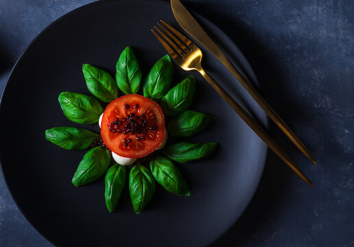 Variation of Caprese salad, slices of fresh ripe tomato, mini mozzarella, leaves of fresh basil and balsamic caviar..Dark background table. Close up, top down view.