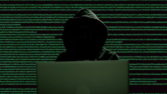 silhouette of a man in a hood working as a hacker at the computer in the dark room at night, hacking the system and laundring secret information.