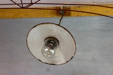 A portrait of an old round metal ceiling lamp hanging from a beam of the ceiling with a light bulb in it. The light is switched off and it is a but rusty.