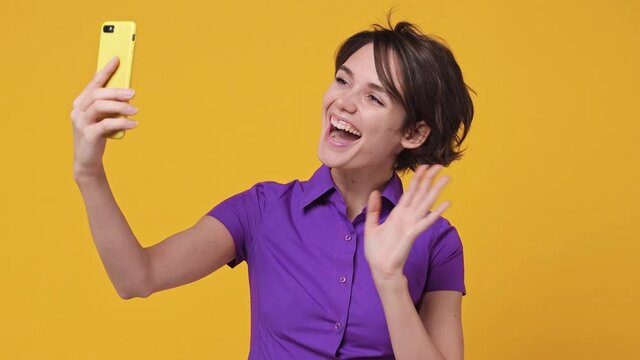 Smiling woman 20s years old in violet shirt get video call use mobile cell phone doing selfie videoconference talk conducting pleasant conversation greet with hand isolated on yellow background studio