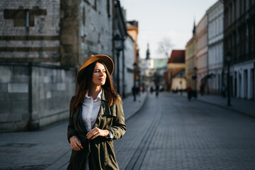 Fototapeta premium Outdoor fashion portrait of elegant, luxury woman wearing beige hat, sunglasses, trendy white shirt, in a green trench coat, walking in street. Copy, empty space for text