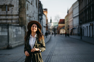 Fototapeta premium Outdoor fashion portrait of elegant, luxury woman wearing beige hat, sunglasses, trendy white shirt, in a green trench coat, walking in street. Copy, empty space for text