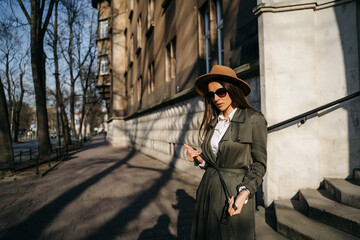 Fototapeta na wymiar Outdoor fashion portrait of elegant, luxury woman wearing beige hat, sunglasses, trendy white shirt, in a green trench coat, walking in street. Copy, empty space for text