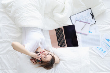 Smiling woman talking on phone and working on laptop while lying on white bed