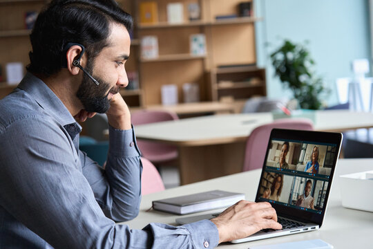 Indian Business Man Wearing Headset Having Virtual Team Meeting On Video Conference Call, Using Laptop Work From Home Office Talking To Diverse People Group In Remote Teamwork Online Distance Chat.