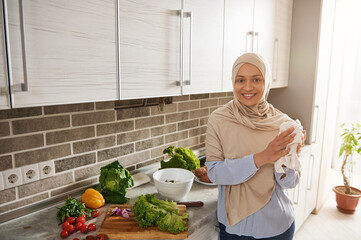 A cheerful Muslim woman in hijab looks at camera and smiles with toothy smile while wiping her hands with a towel standing against the background of her kitchen.