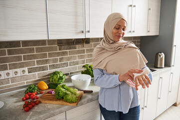 A charming Muslim woman in hijab wiping her hands with a towel standing against the background of her kitchen.