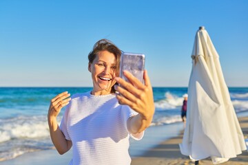Happy middle-aged woman talking on smartphone using video call, on beach