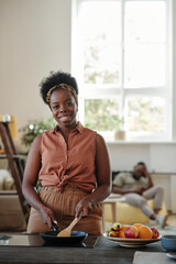 Young smiling woman of African ethnicity looking at you while standing by electric stove