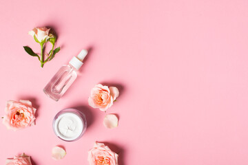 Bottles of serum and cream tube with rose flowers flat lay on pink background