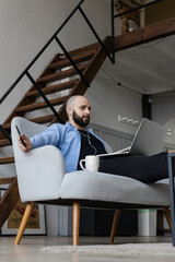 A guy in a blue shirt and black jeans is sitting on the couch and working at a computer. The concept of freelance work