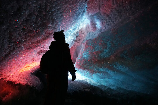 A guy in an ice cave with a lantern light. The caver descended into the ice cave. Snow stalactites and ice walls. In some places there are stones. Colored lantern beams. Bogdanovich Glacier, Almaty