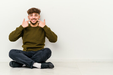Young Moroccan man sitting on the floor isolated on white background smiles, pointing fingers at mouth.