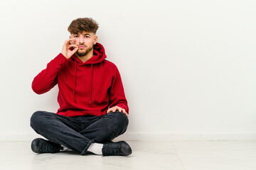 Young Moroccan man sitting on the floor isolated on white background with fingers on lips keeping a secret.