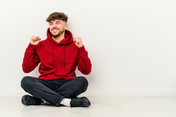 Young Moroccan man sitting on the floor isolated on white background celebrating a victory, passion and enthusiasm, happy expression.