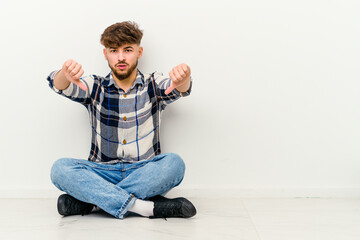 Young Moroccan man sitting on the floor isolated on white background showing thumb down and expressing dislike.