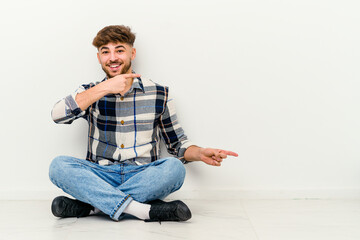 Young Moroccan man sitting on the floor isolated on white background excited pointing with forefingers away.