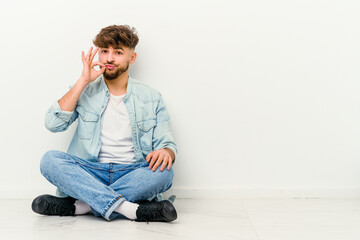 Young Moroccan man sitting on the floor isolated on white background with fingers on lips keeping a secret.