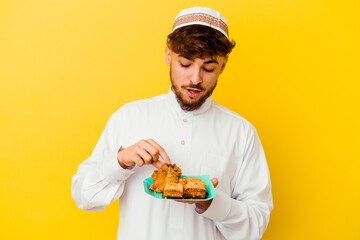 Young Moroccan man wearing the typical arabic costume eating Arabian sweets isolated on yellow background