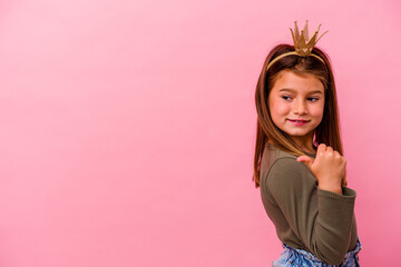 Little princess girl with crown isolated on pink background points with thumb finger away, laughing and carefree.